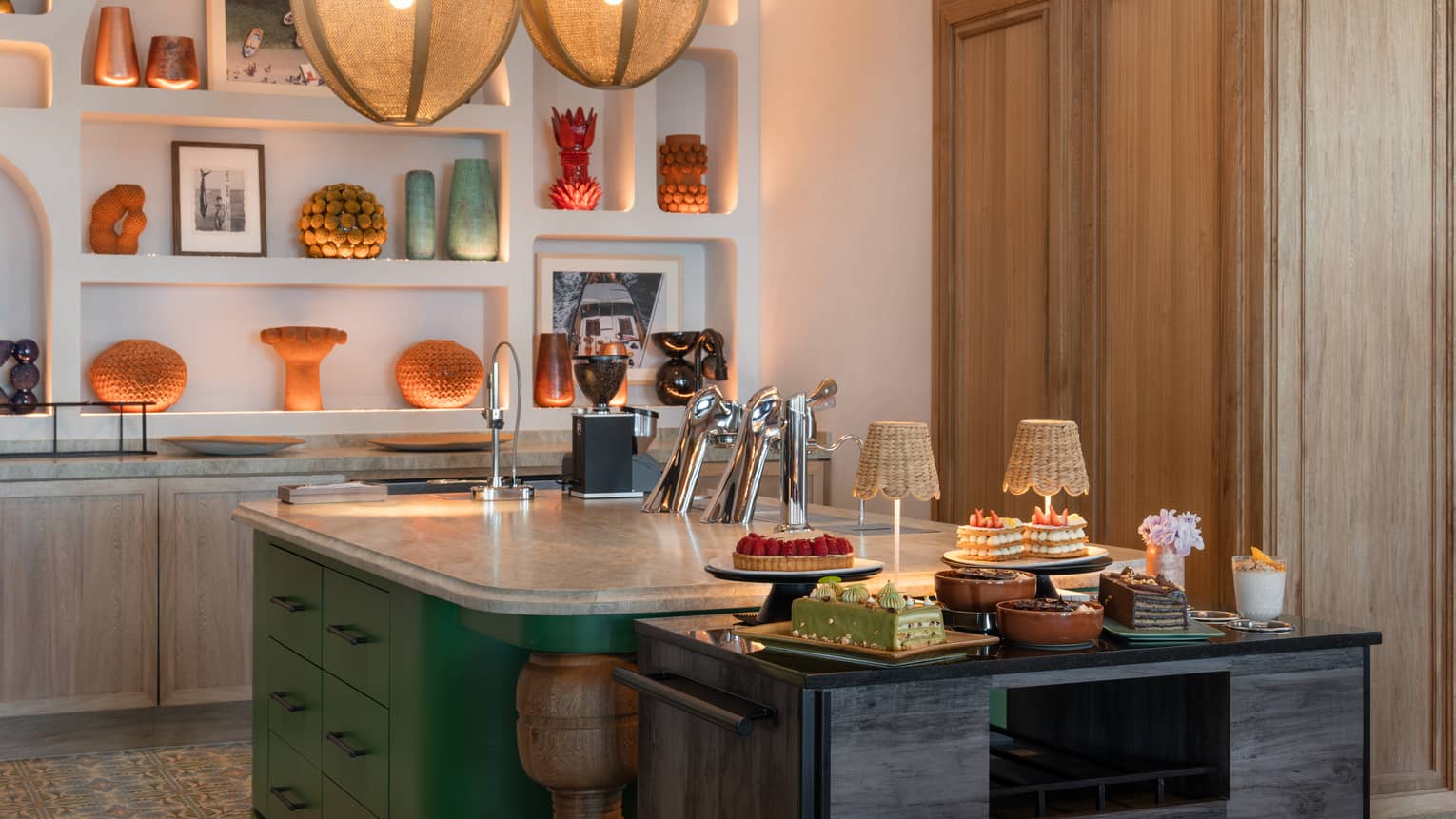 Large kitchen island with marble top and green cabinetry next to a black table holding a variety of pastries