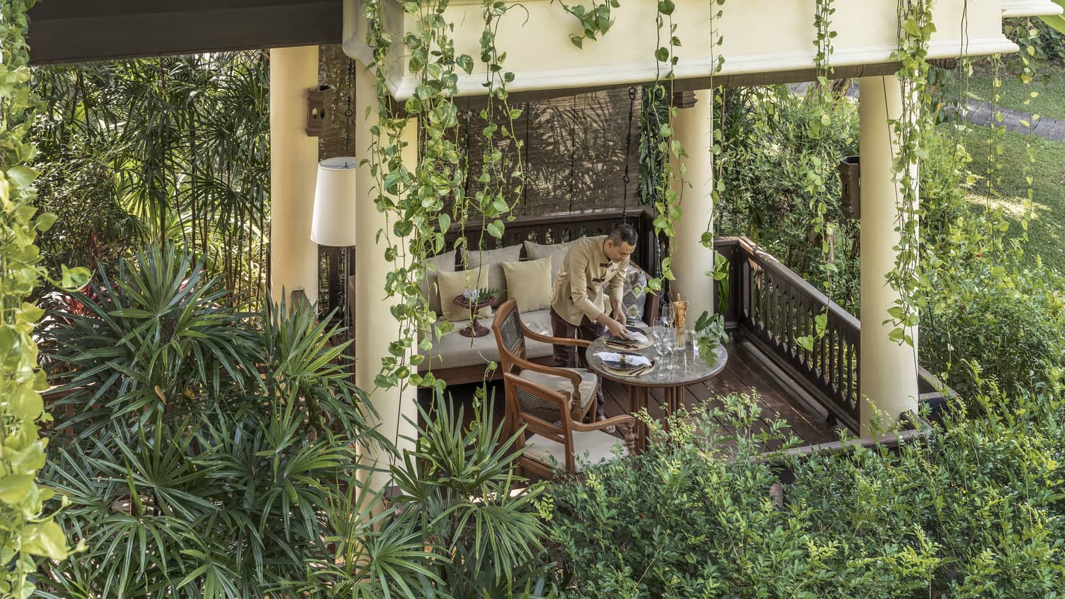 Person setting a dining table in a covered outdoor seating area surrounded by hanging plants and lush greenery