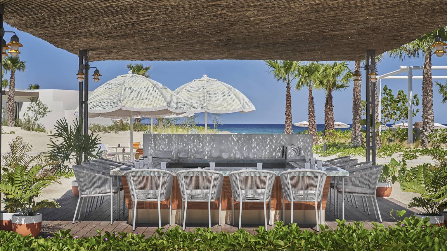 An open air dining area under a pavilion, surrounded by greenery and plantings, with ocean views in the background