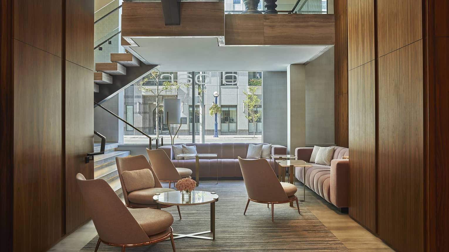 A lounge area with upholstered sofas and chairs, marble tables and a staircase in front of a large window