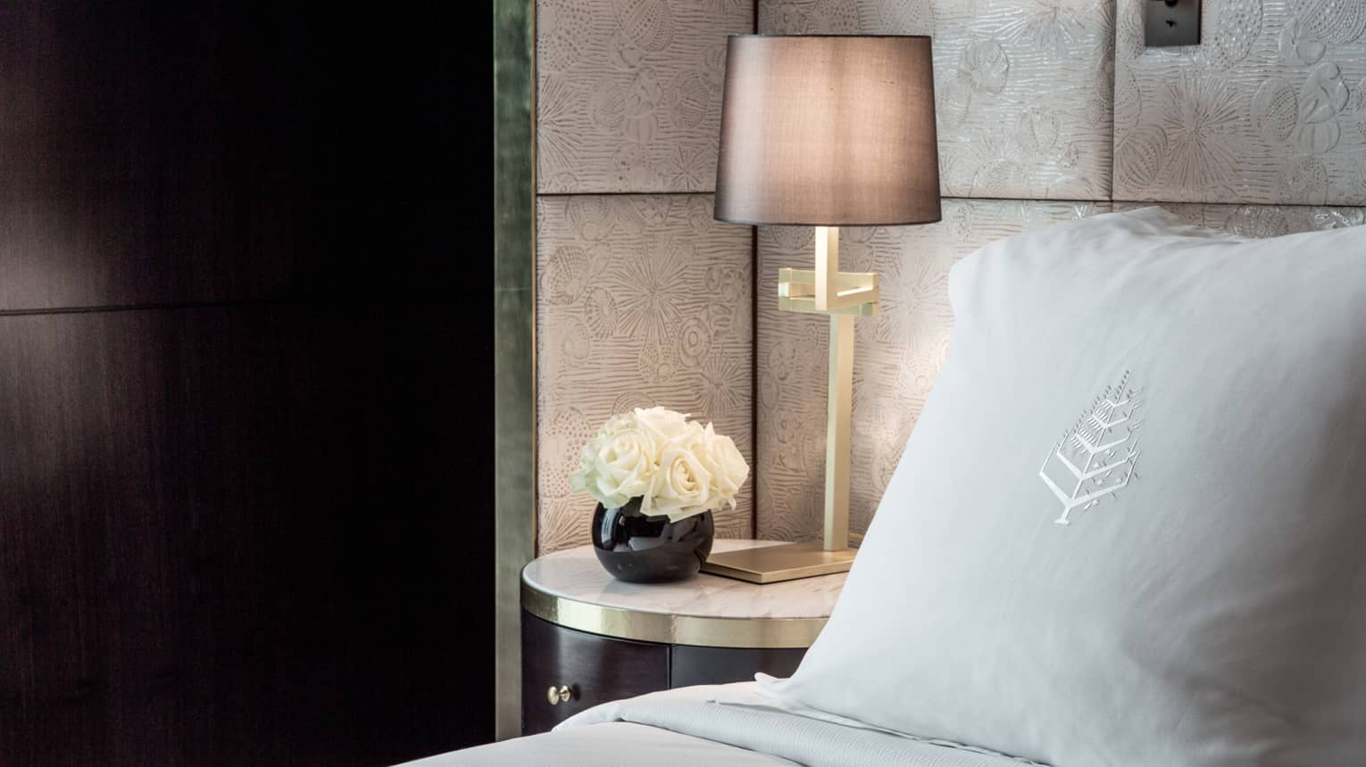 Premier room, close-up of pillow with Four Seasons logo, white roses in round black vase and bedside reading lamp