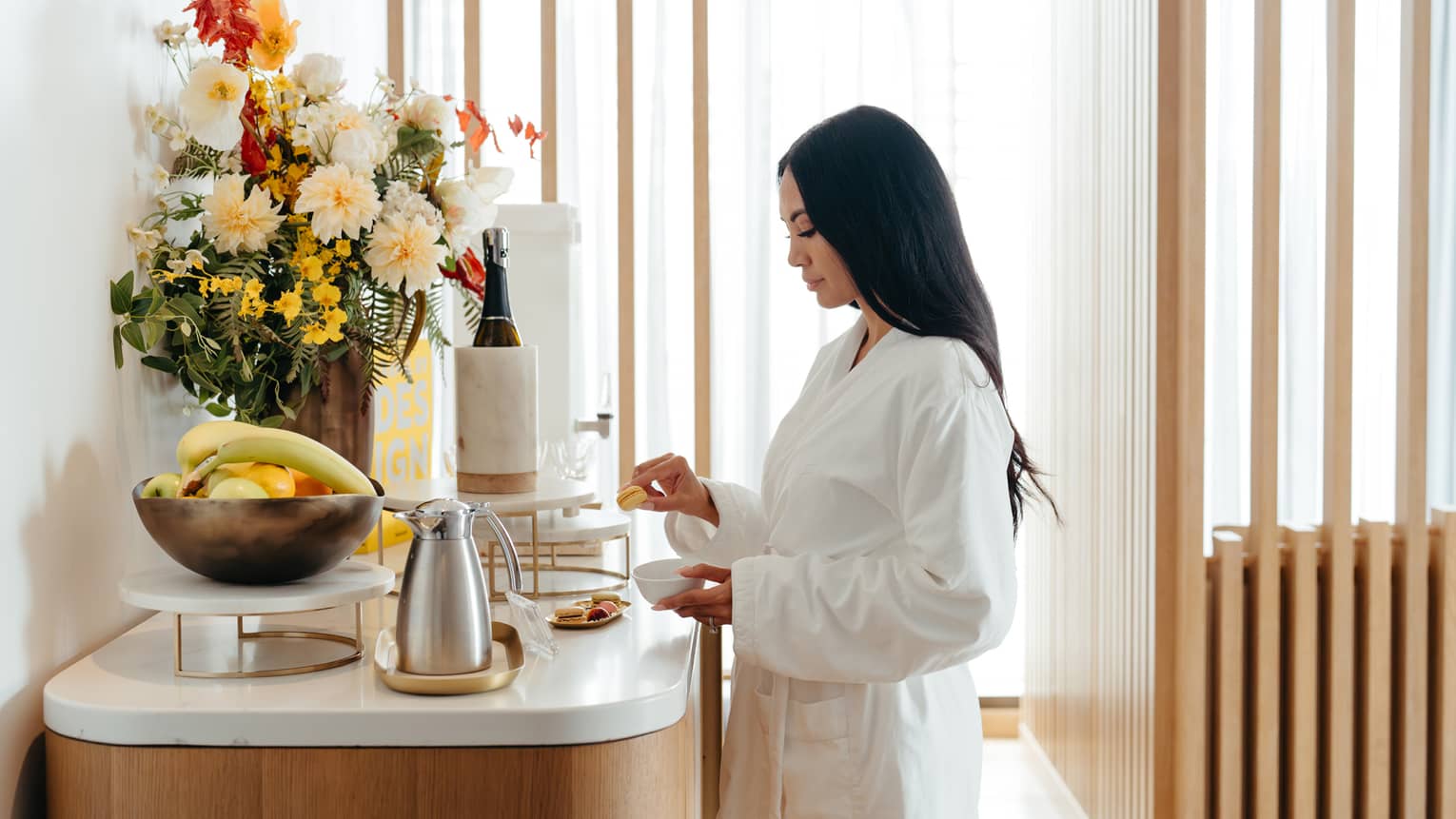 A guest in a white robe stands in a light-filled interior space in front of a counter holding a floral arrangement, a bowl of fresh fruit, a plate of small food items, a carafe and other beverage items.