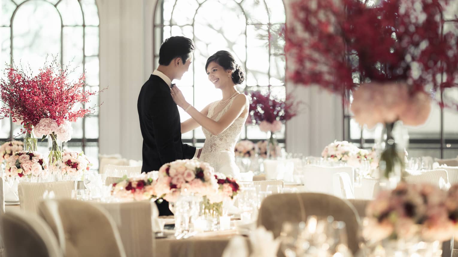 Woman in wedding dress adjusts groom's bow tie in light filled banquet room with pink flowers