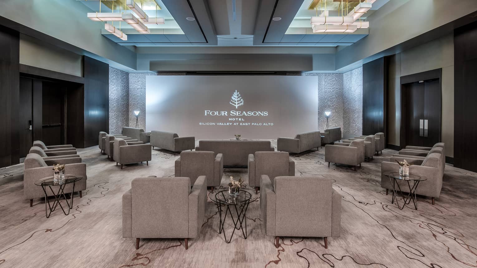Gray plush chairs are angled towards a large projector in an indoor carpeted event space