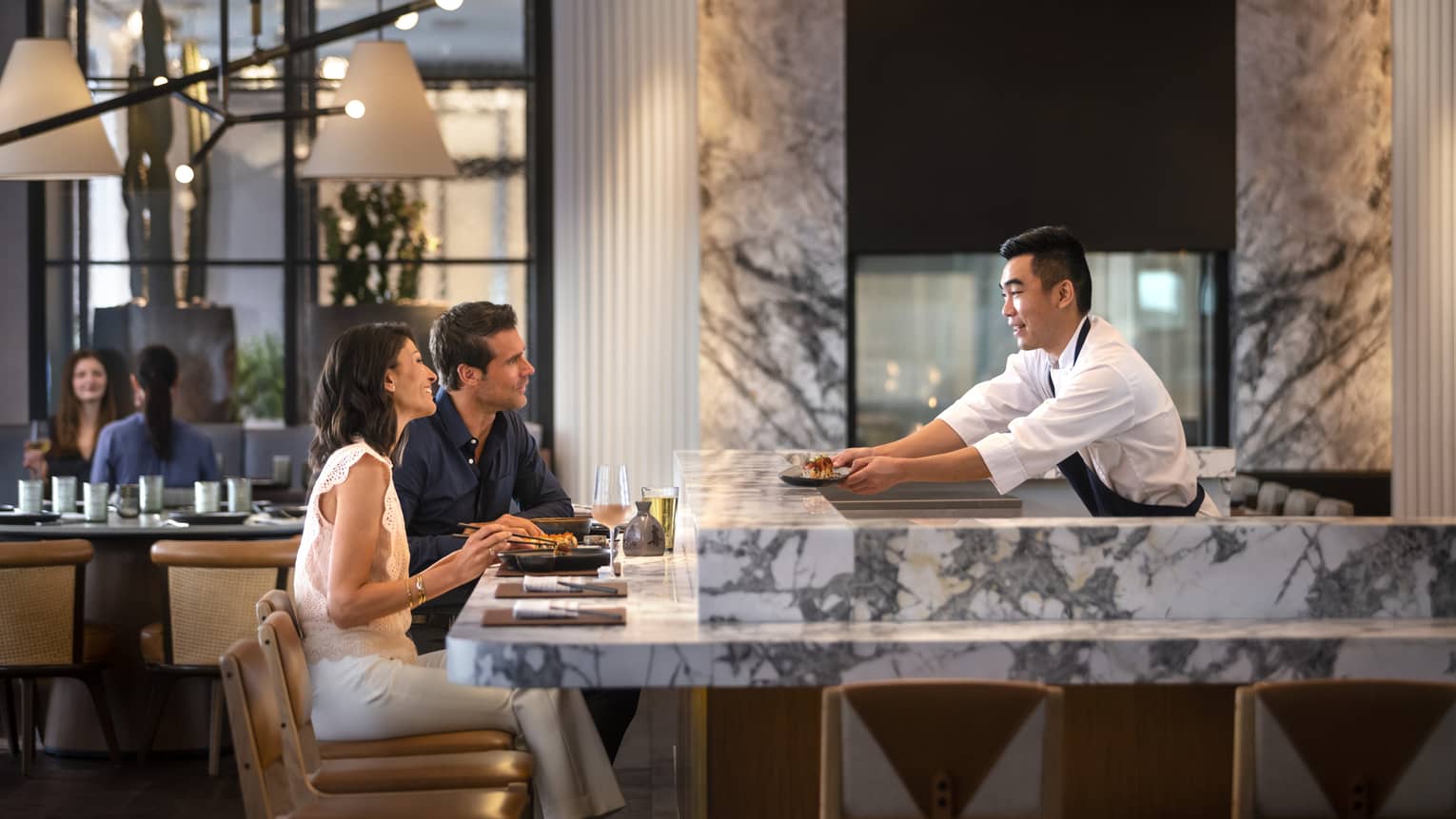 A couple sitting at a bar being served sushi.