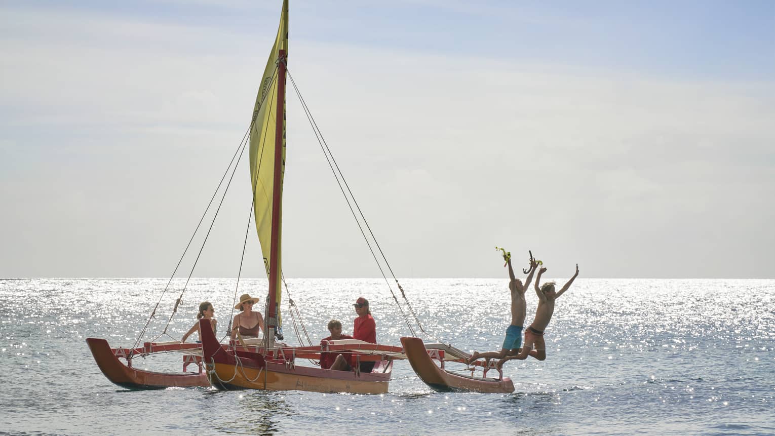A family floats on an outrigger canoe as two people jump off the boat into the water