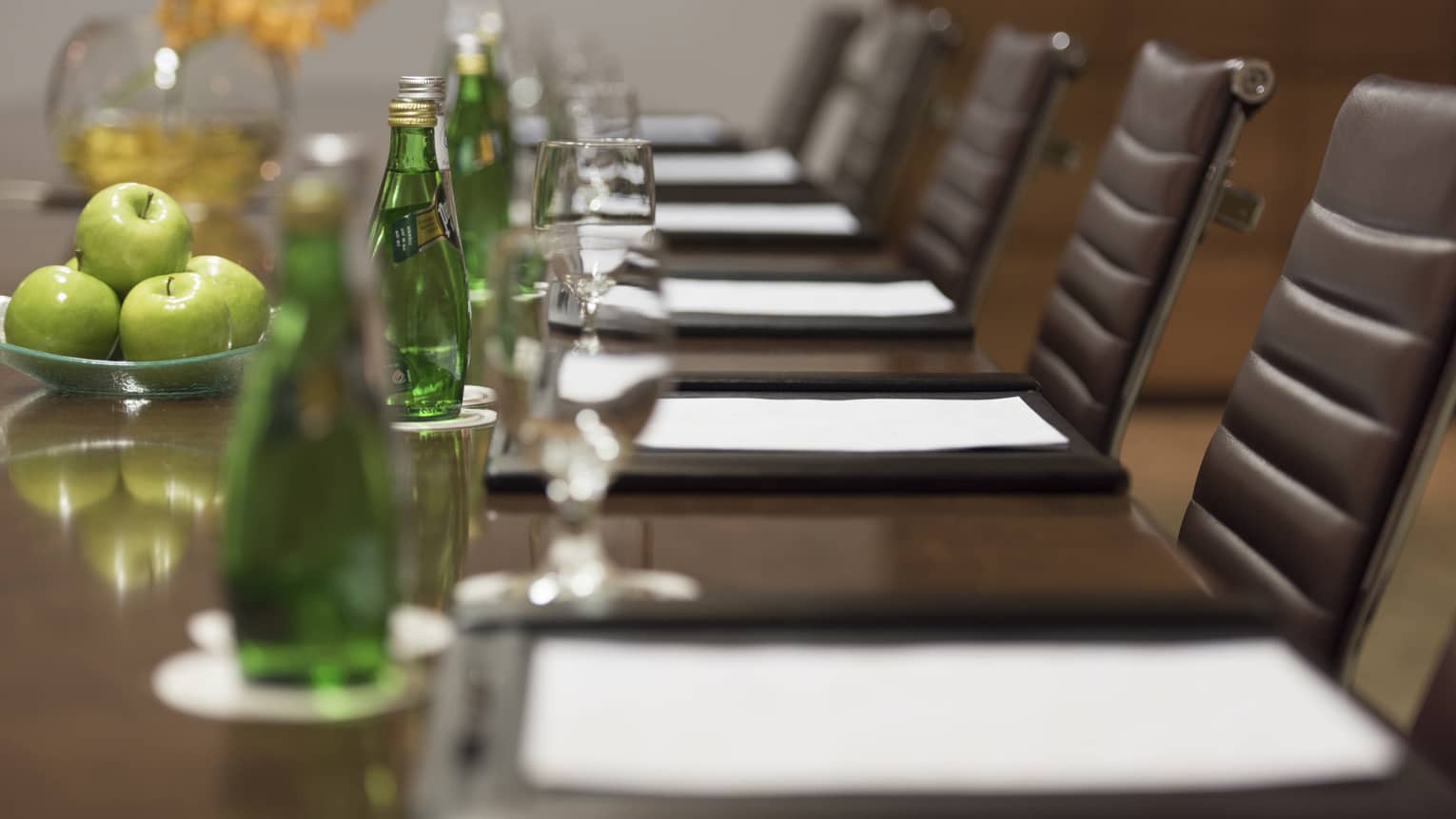 Close-up of business meeting table row of chairs, paper agendas, green glass bottles, apples
