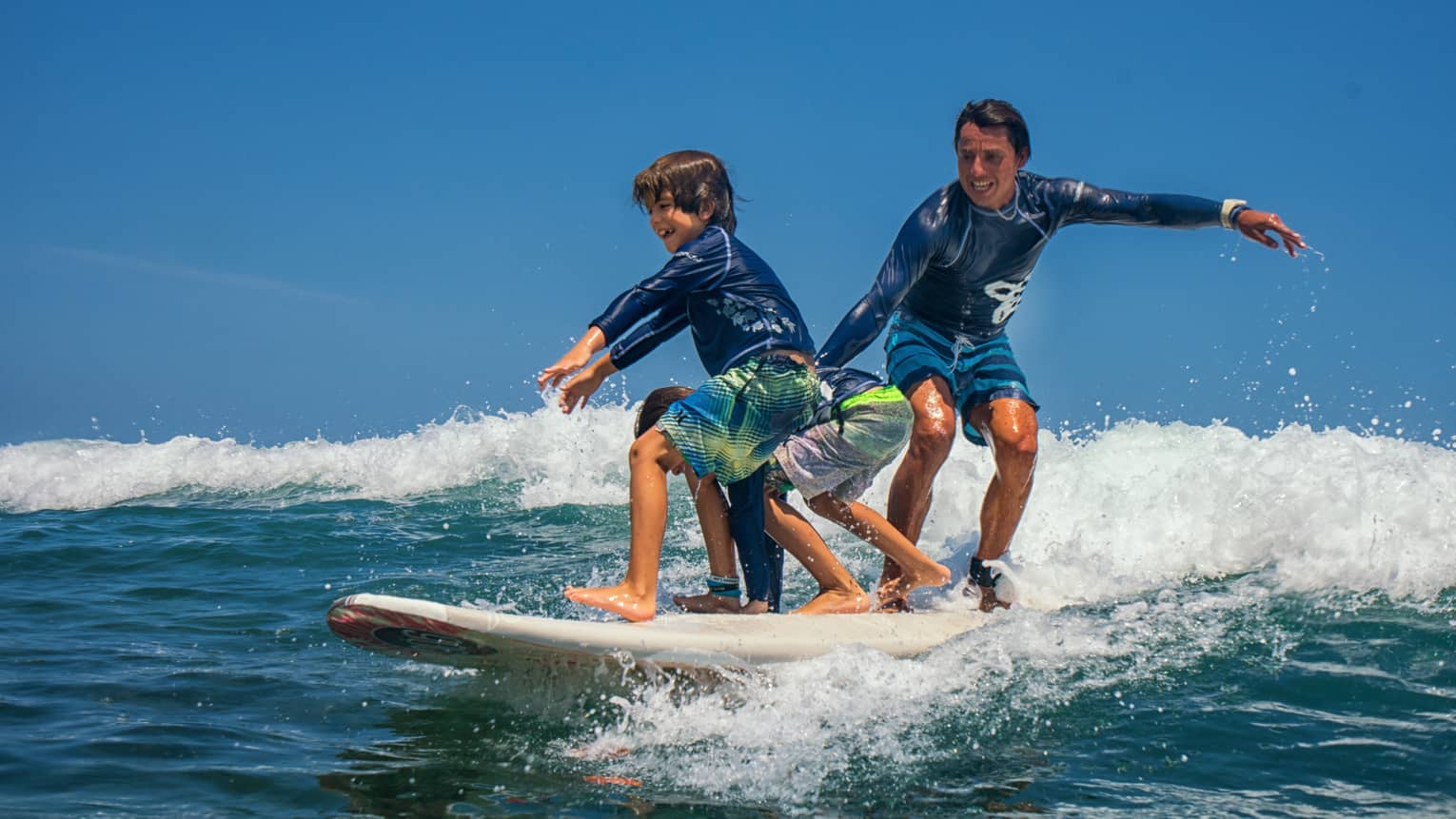 Adult and two children share a surfboard, laughing as they balance on a foamy wave. 