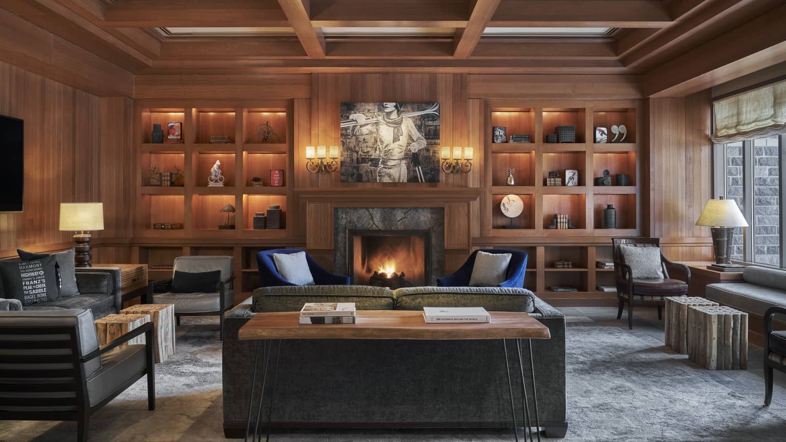 An oak library with several illuminated shelves, and a roaring fire in the stone fireplace.
