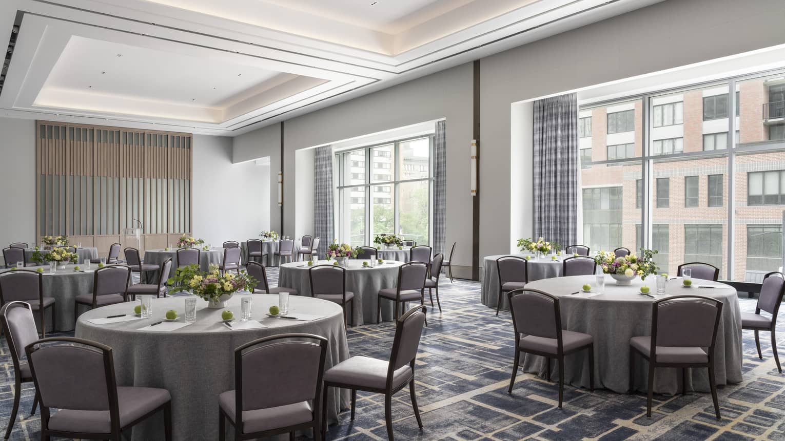 A ballroom with round tables covered in grey table cloth and yellow flower, with large windows in the room.