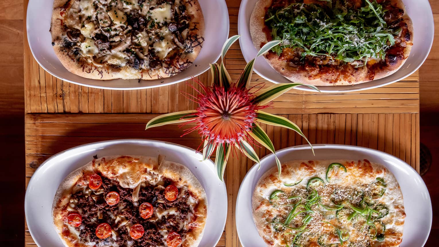 Four different pizzas including tomato, mushroom, cheese, greens, and more on a wood table.