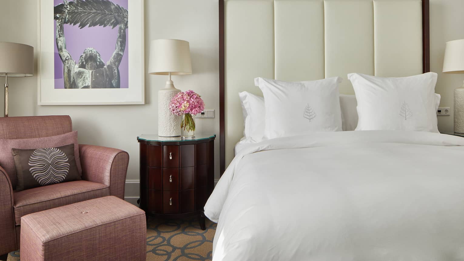 A room with a bed with white sheets, a chairs, side table and art on a wall.