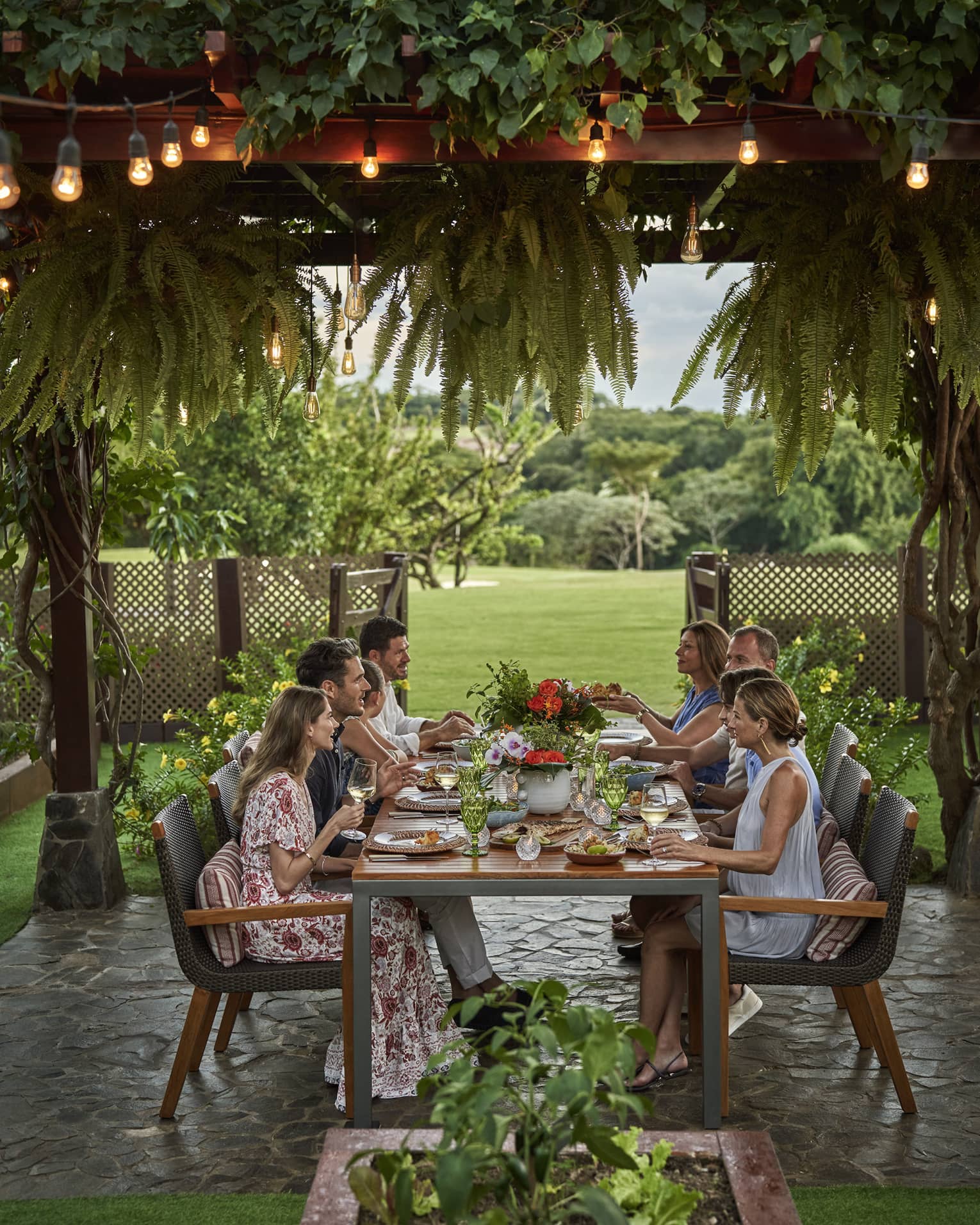 Eight people sit around a long dining table set up outside beneath a canopy of greenery and string lights
