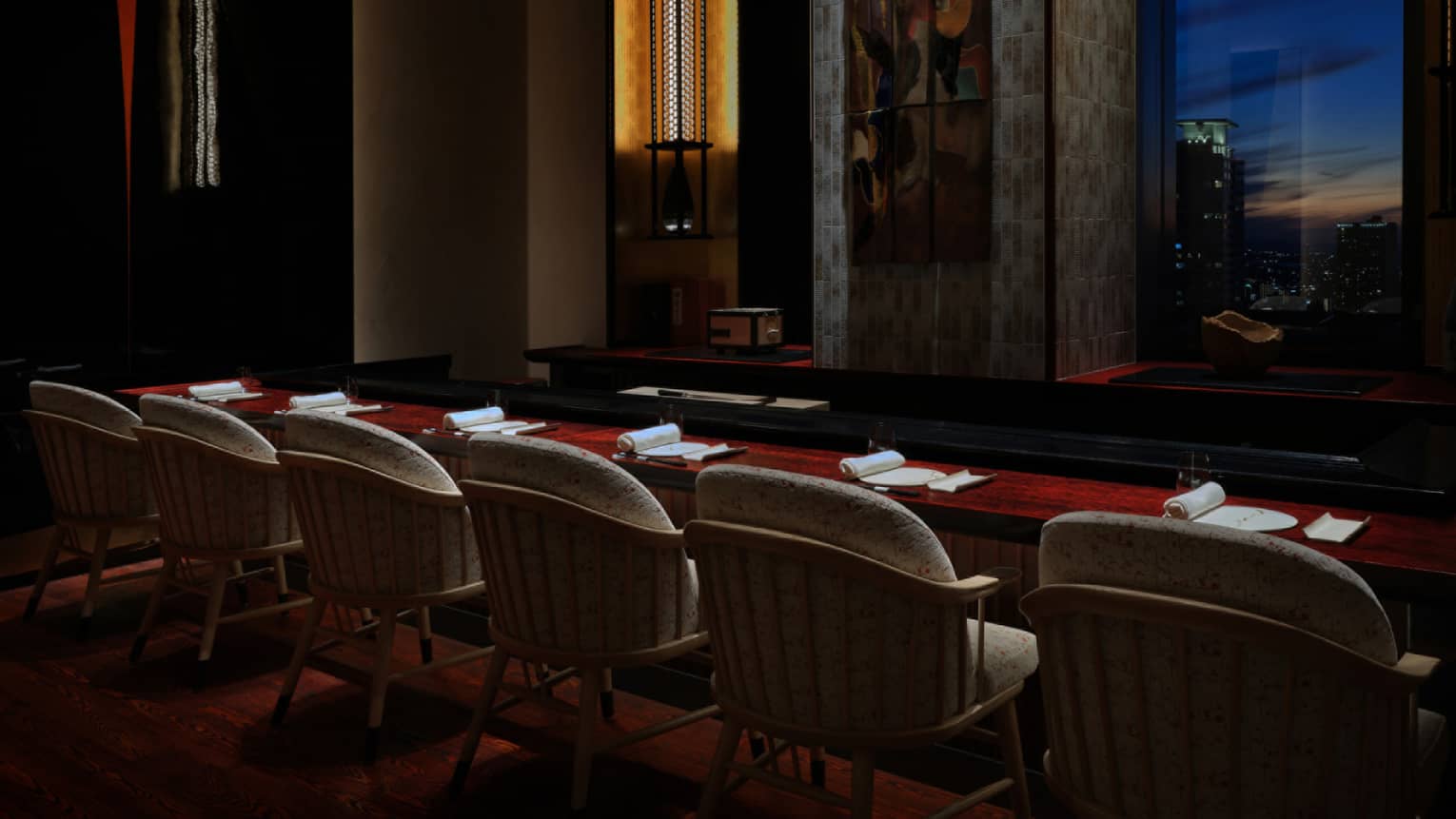 In a dark room, a long mahogany table with six white place settings faces a window looking out at the city in the evening.