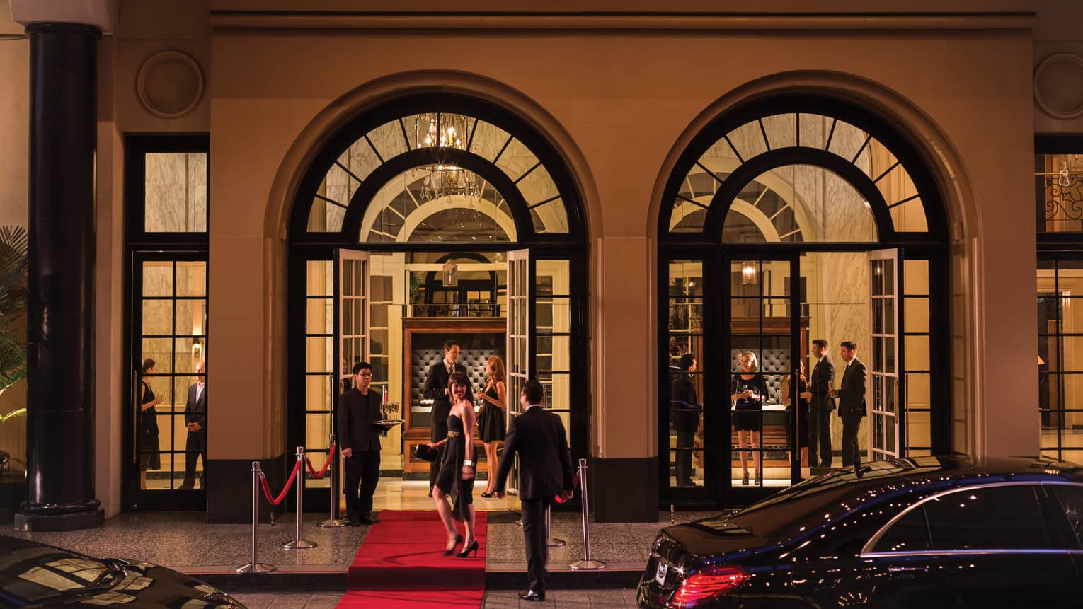 Party entrance, smiling woman wearing black cocktail dress stands on red carpet under arched doorways