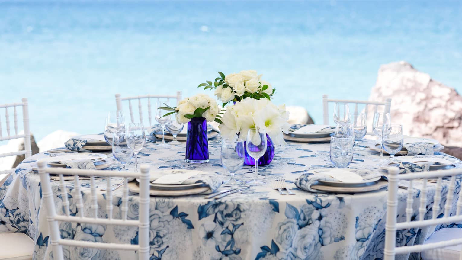 Blue and white round table formally set, five white chairs, white floral centrepieces, on beach