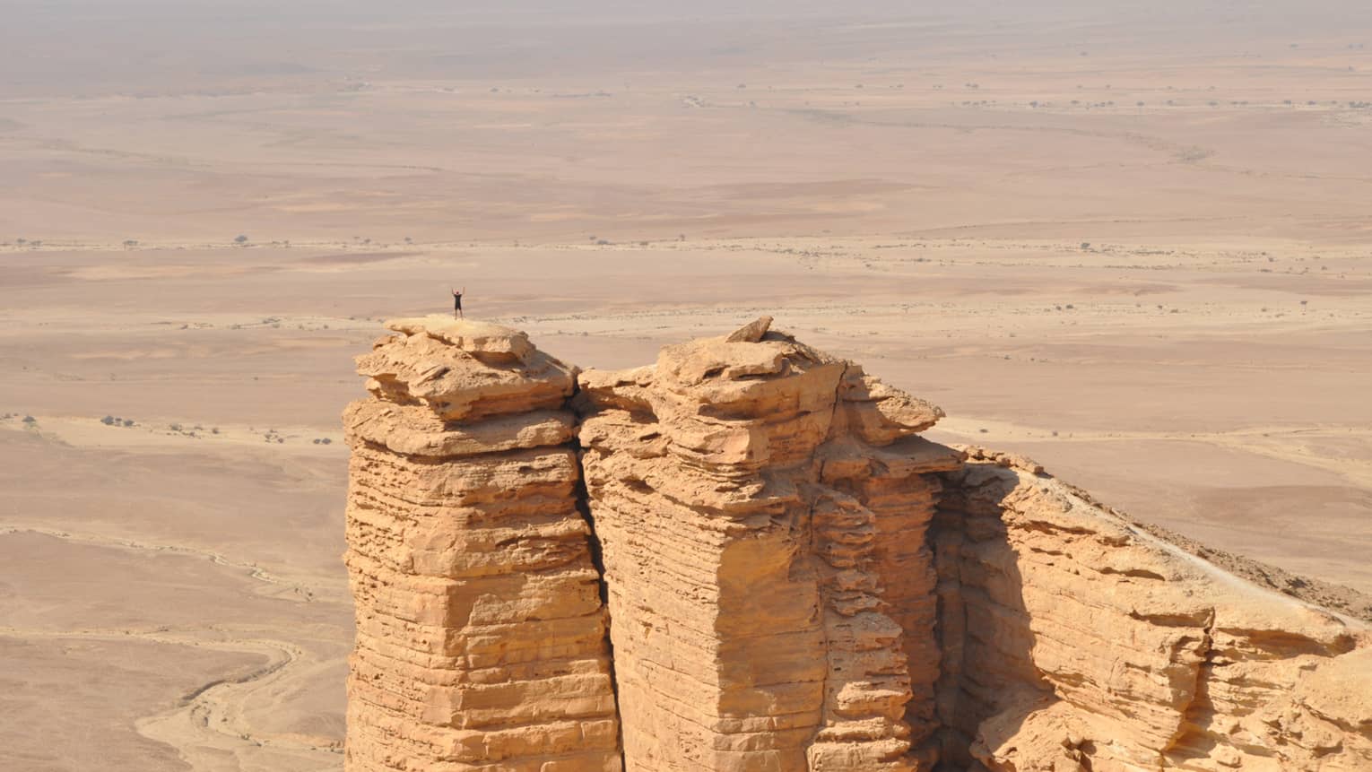 An aerial view of a giant golden cliff surrounded by a dry desert and a small silhouette of a person standing on top of it.