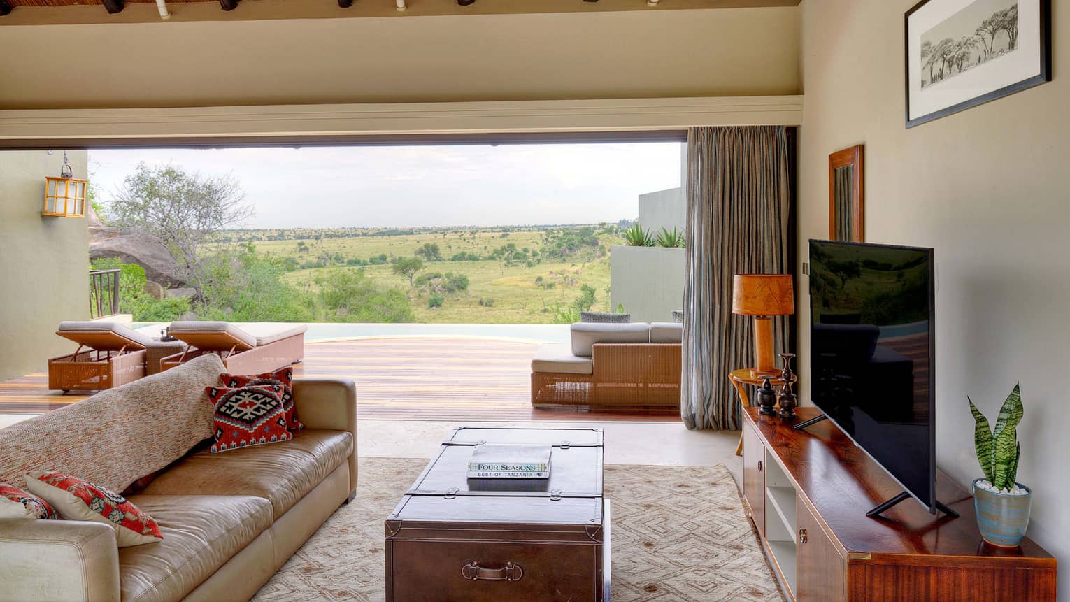 Villa seating area with beige leather sofa, trunk coffee table, flat-screen TV on console and expansive plains views outside open wall to terrace