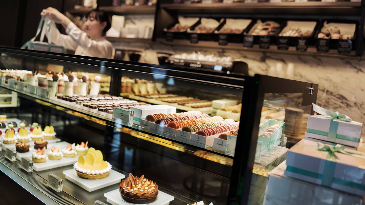 Pastry chef ties bow on box behind large glass counter with pastries, desserts