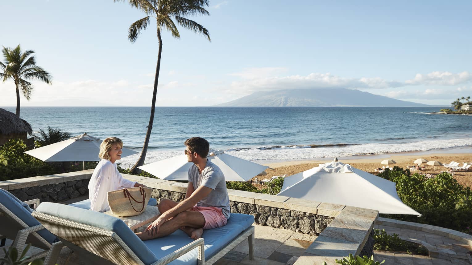 Man in shorts and smiling woman in beach shirt sit on edge of lounge chair, beach and ocean in background