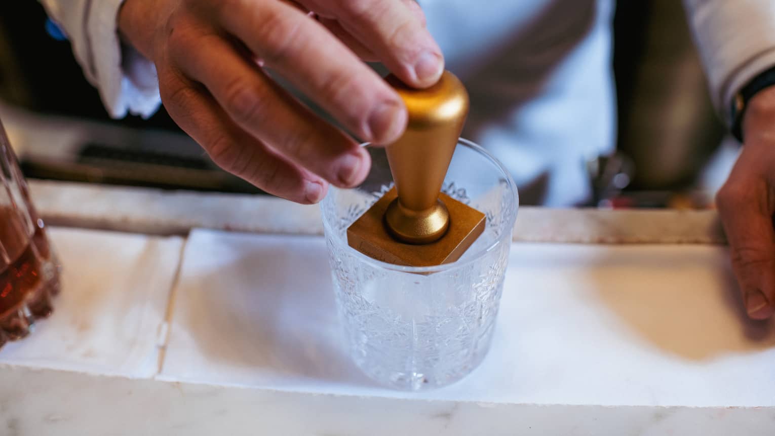 Close-up of a hand embossing an ice cube using a brass ice stamp in a clear cut-crystal lowball cocktail tumbler.