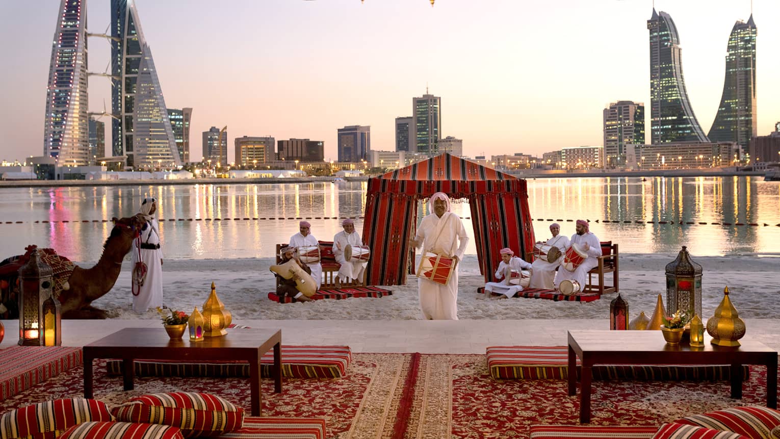 Band plays on white sand beach by lounge at sunset, city skyline across water