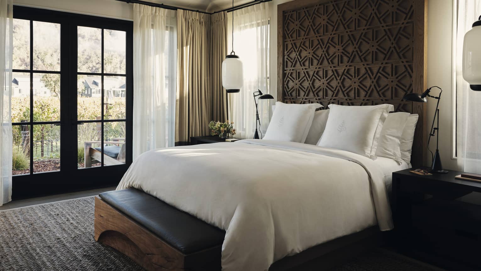 Luxury residential villa bedroom with a king bed, carved wood headboard, bench, nightstands, pendant lights and large windows with a vineyard view