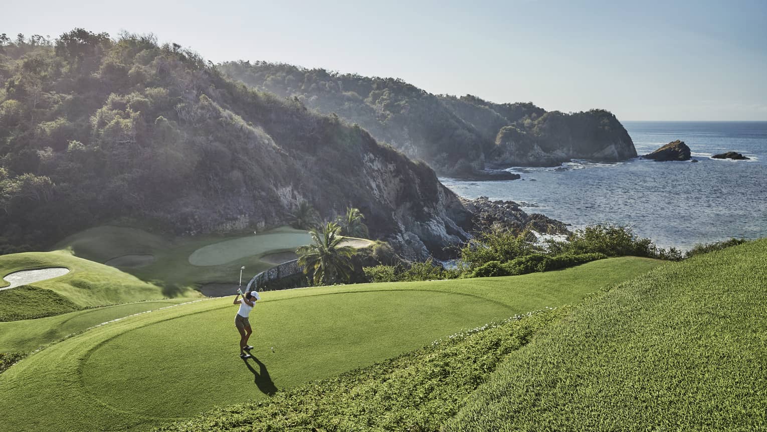 Golfer in mid-backswing, ready to strike toward a distant green at the base of an immense forested hill bordering the ocean. 