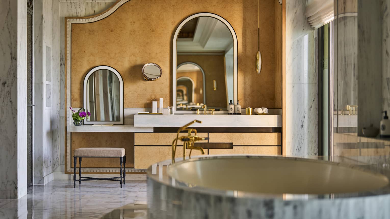,Large suite bathroom with free-standing oval marble bathtub, marble walls and a vanity area with two seats and arched mirrors