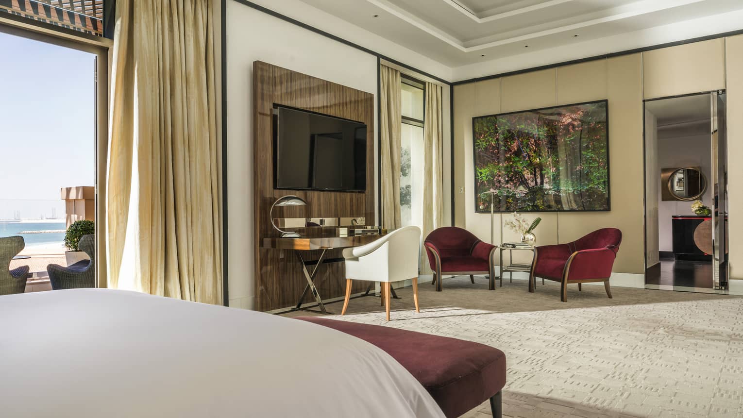 Hotel suite bedroom with large bed, seating area, desk and glass doors that open to a private balcony overlooking the beach
