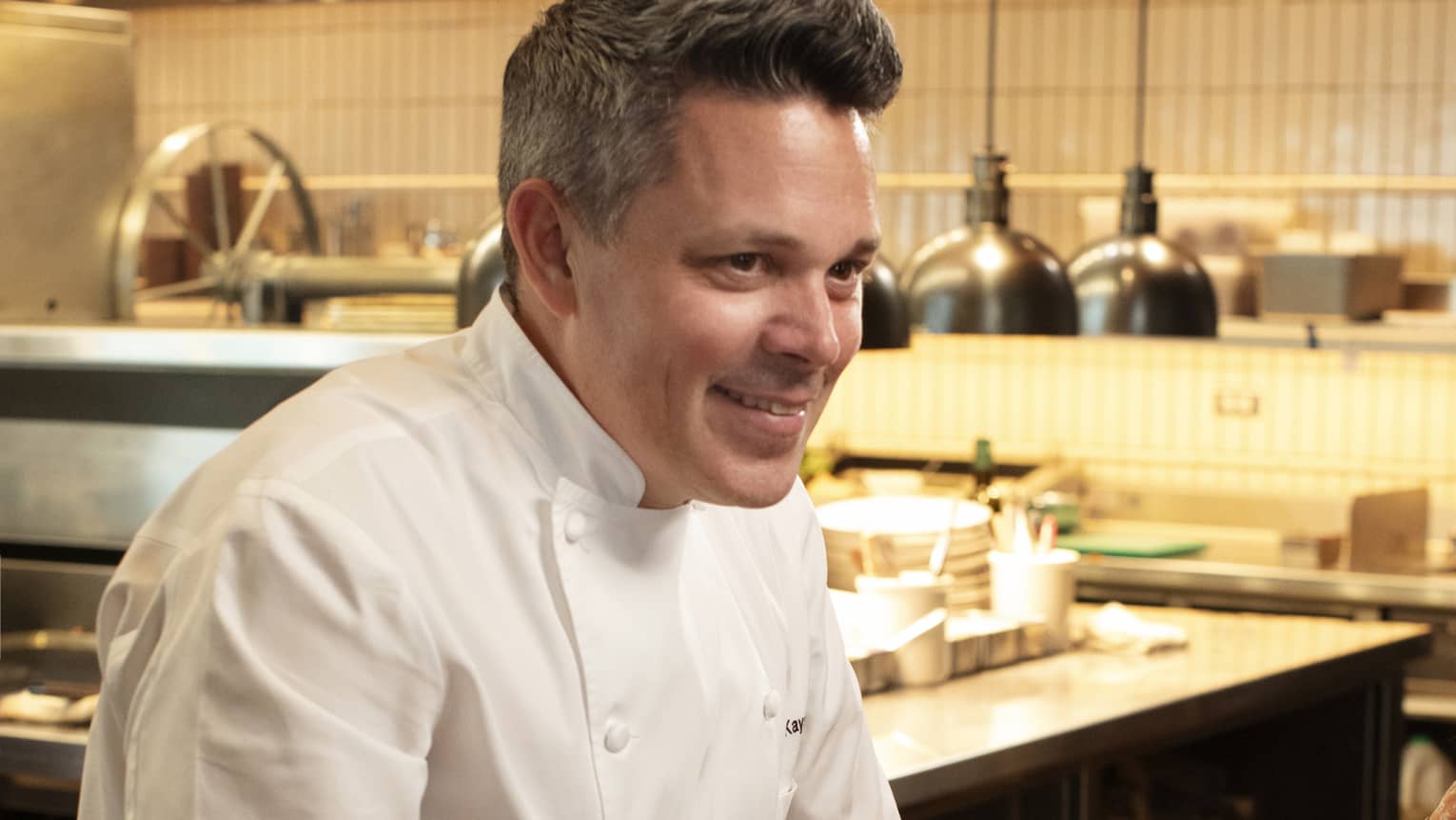 Chef standing beside a dining table in the kitchen talking to guests