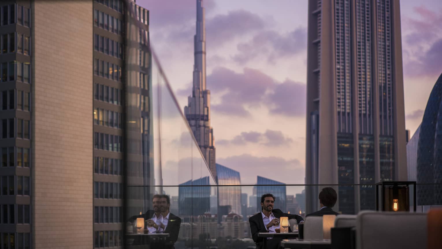 Men in suits on rooftop patio in front of glass balcony wall overlooking city at sunset