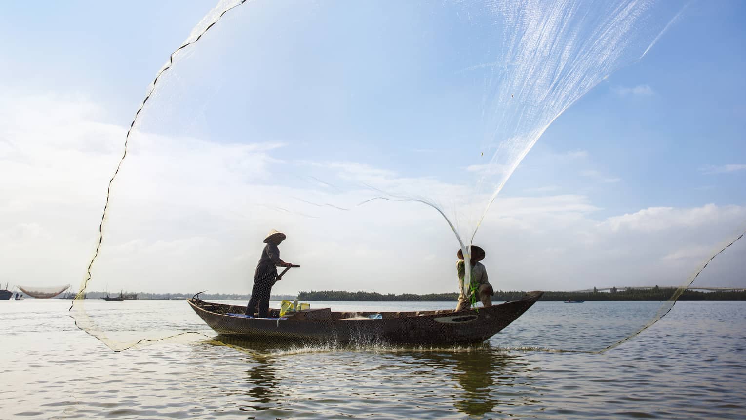 Silhouettes of two people wearing hats in small boat standing, casting large nets 