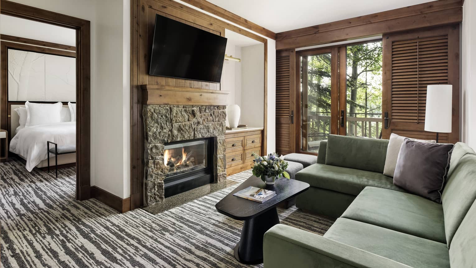 Luxury resort suite living area with a green sectional sofa, stone fireplace, mounted TV and balcony access, adjacent to a bedroom with a king bed