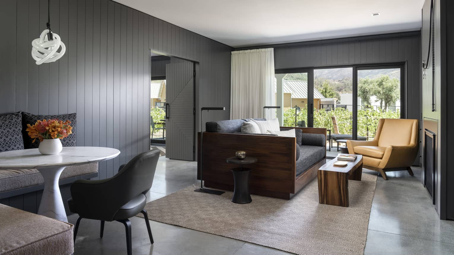 Resort suite living area with a gray sofa, tan armchair, wooden coffee table, small dining area and large windows overlooking greenery