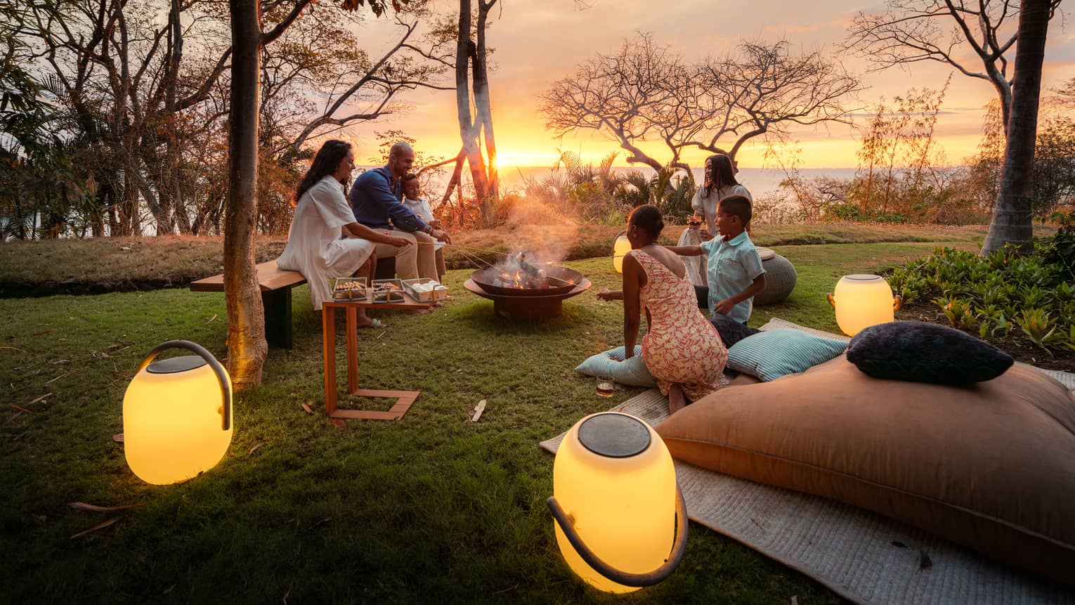 Family of six has a picnic on a private lawn, all seated around a bonfire with lanterns set around them and the sun setting behind them over the ocean
