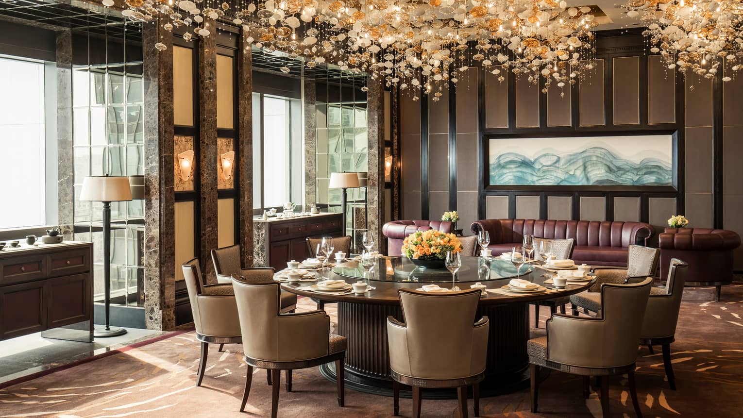 Jin House large round dining table by burgundy leather sofas, under gold, glass lights