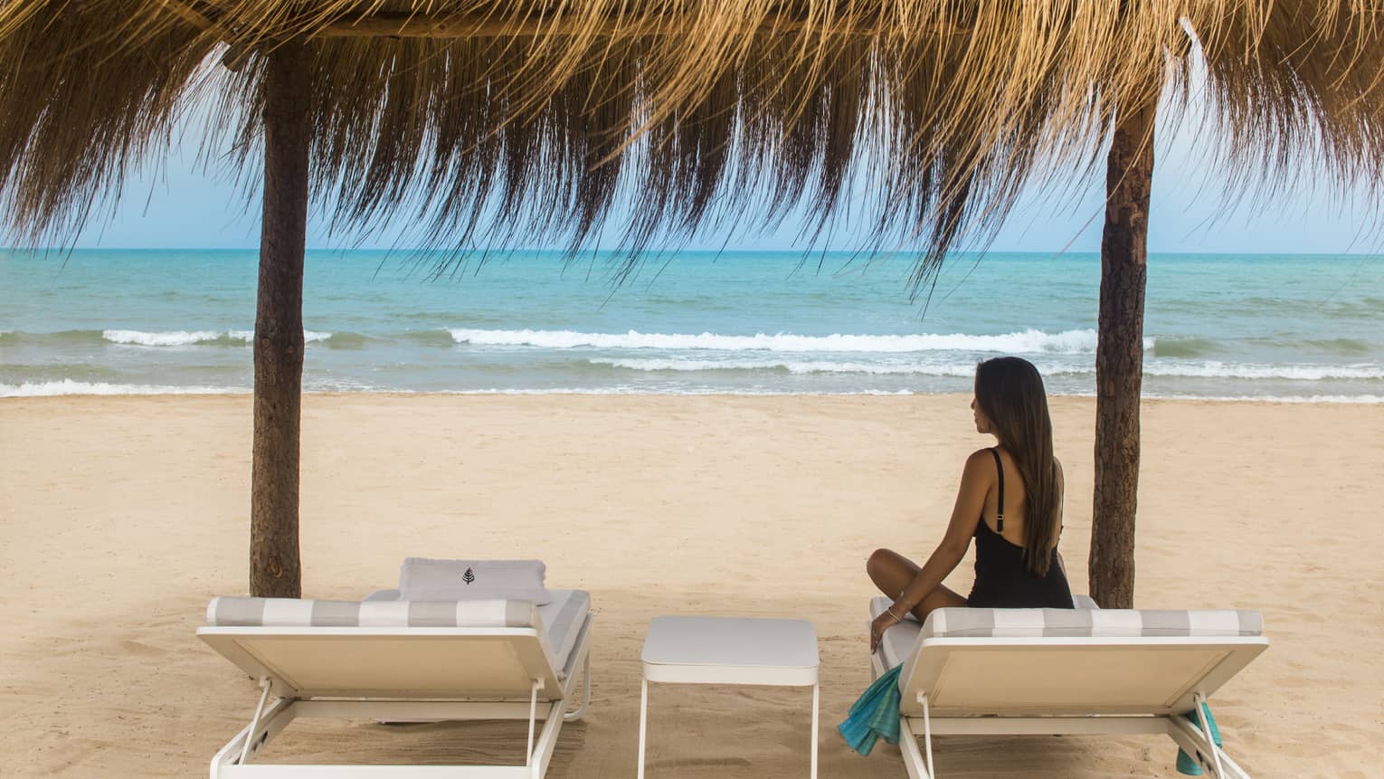 Long-haired woman in black swimsuit sits on lounge chair under grass-roofed beach cabana at Four Seasons Hotel Tunis