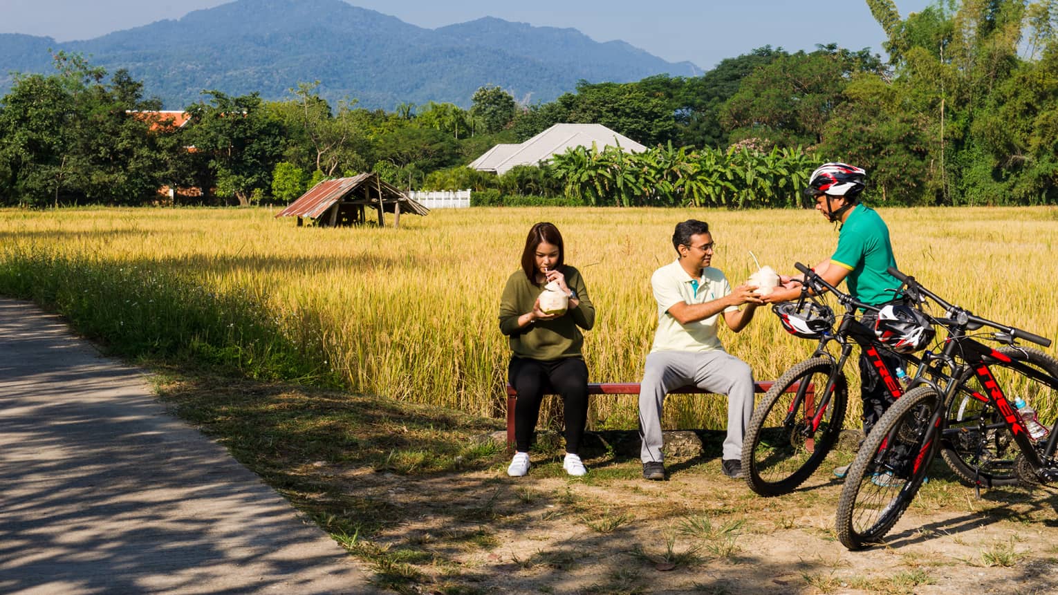 Three mountain bike riders stopped for a rest in a clearing, two seated as a helmeted rider hands a coconut shell cup to one.