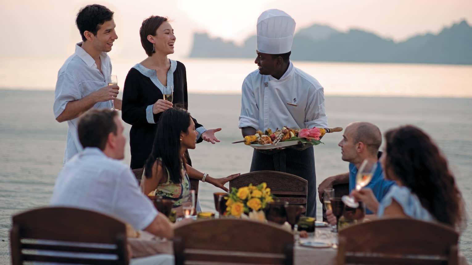 Chef presents platter of grilled meats to group of friends at beach dining table
