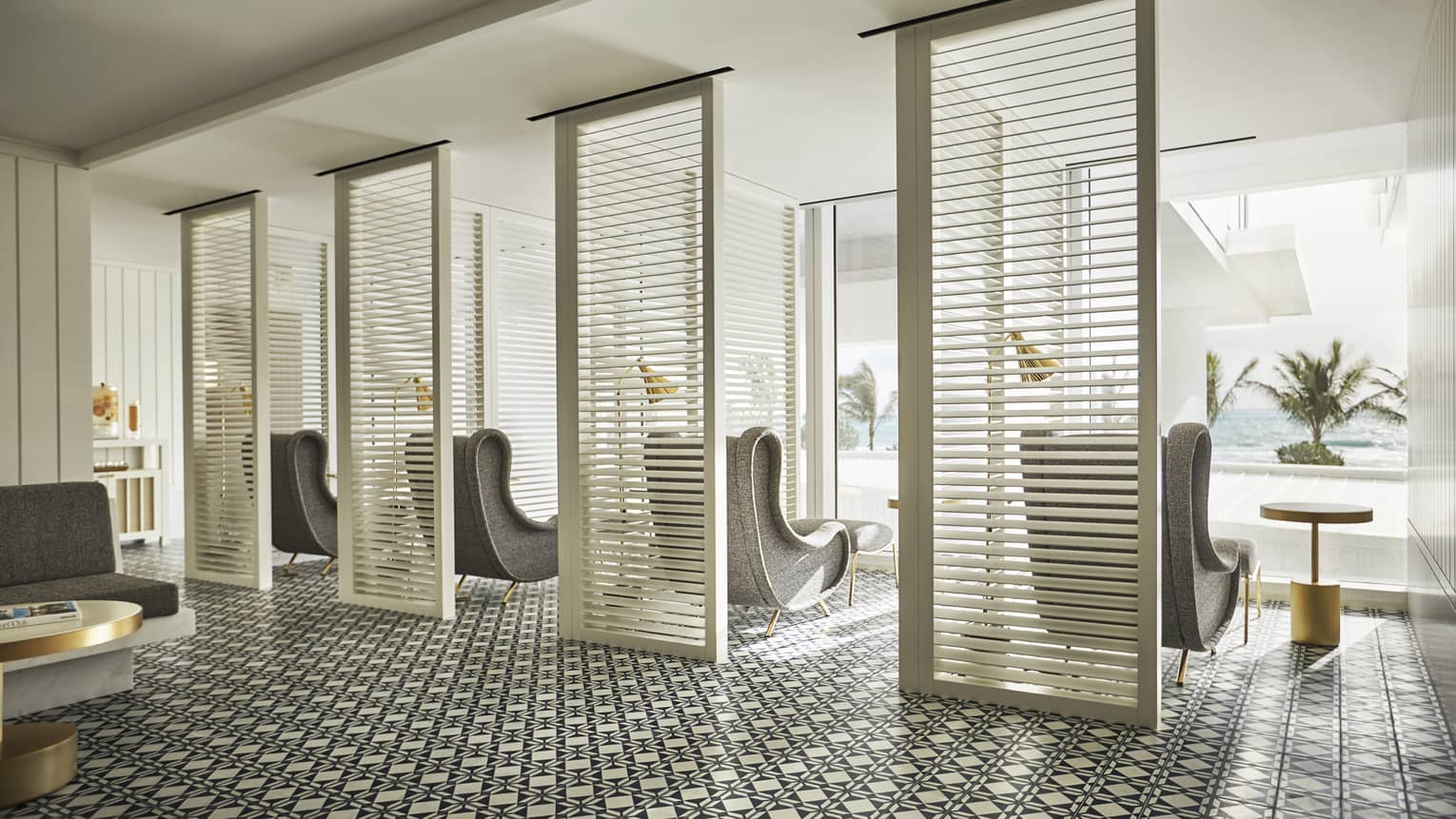 Luxury spa area with patterned tile flooring, partitioned seating space, gray armchairs and floor-to-ceiling windows overlooking ocean and palm trees
