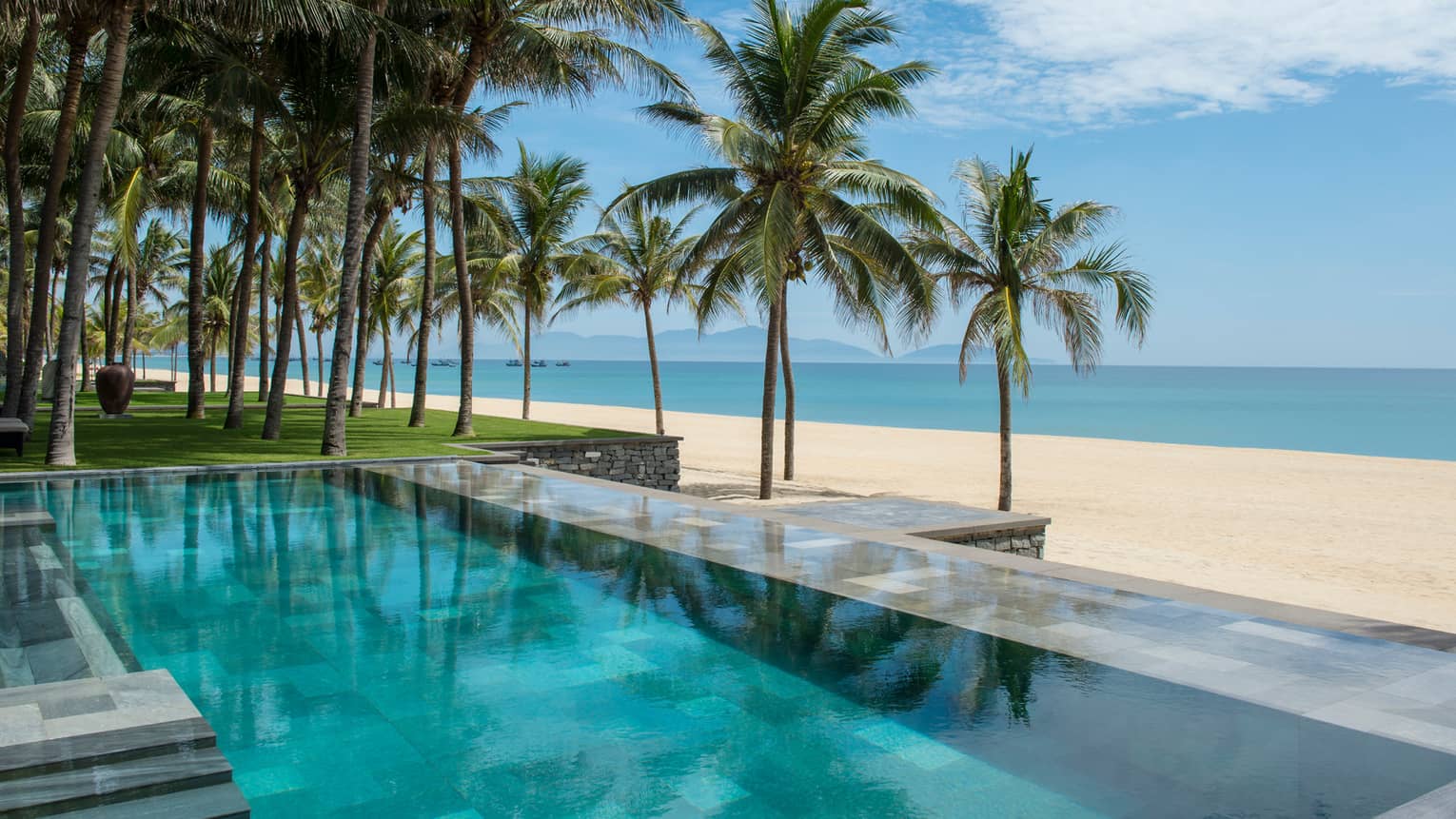 Tall palm trees between stone outdoor swimming pool and white sand beach