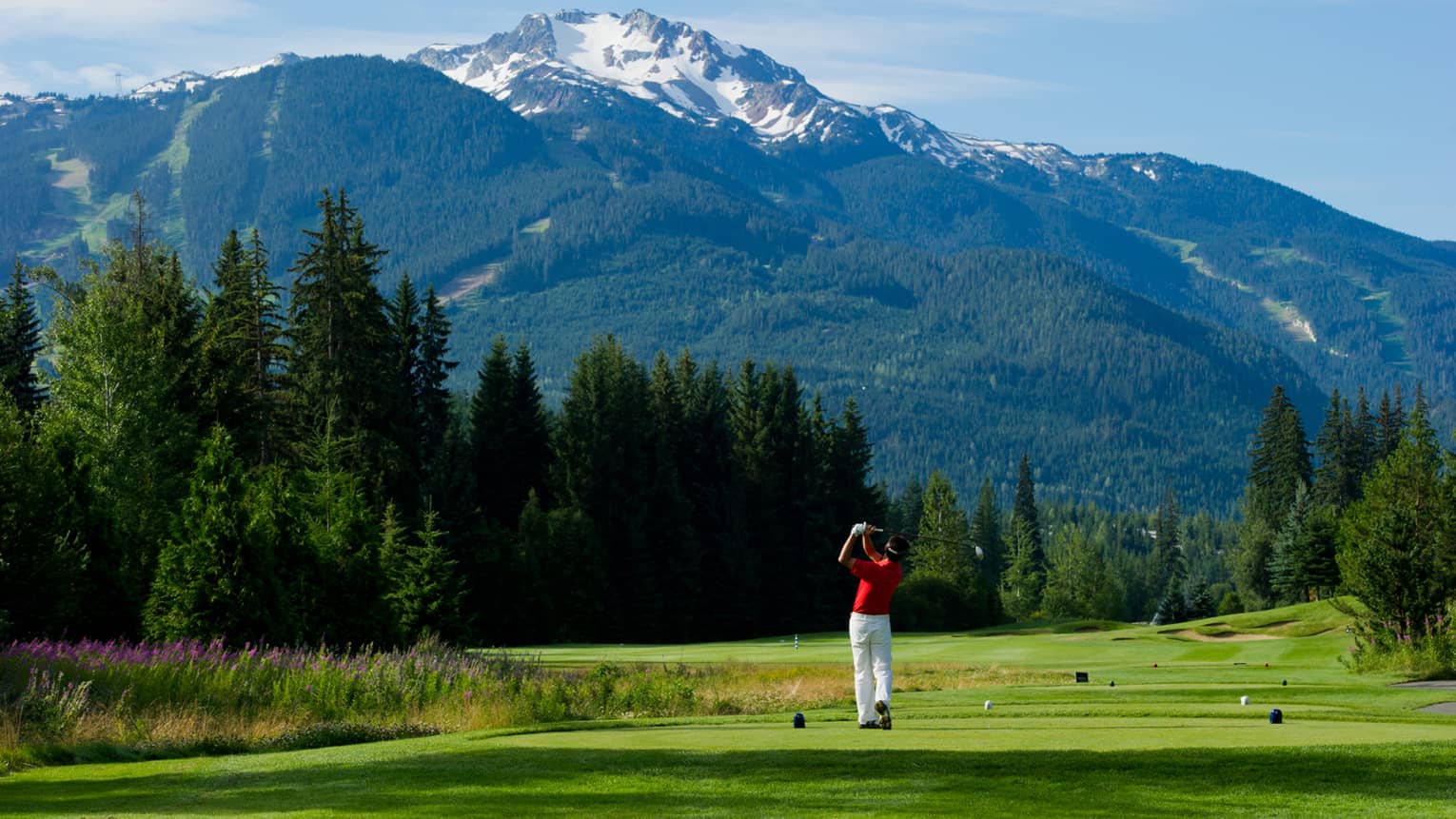 Man wearing red golf shirt swings club on green golf course surrounded by snow-capped mountains