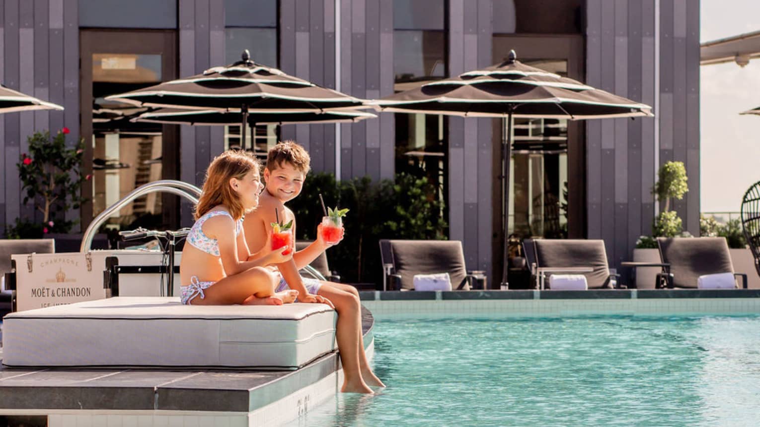 A young boy and girl sitting on the edge of a pool outside.