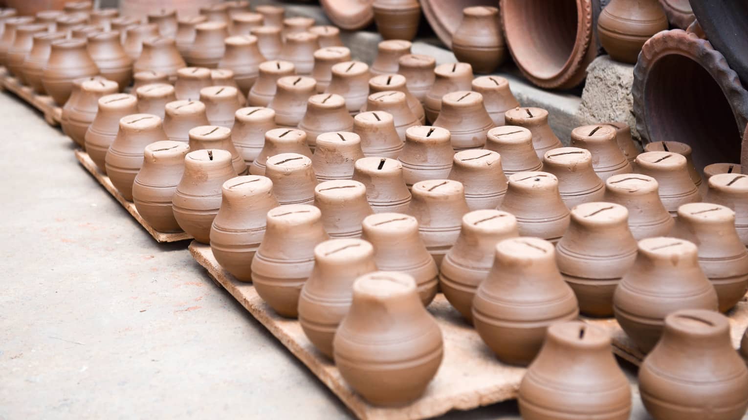 A collection of small, brown clay piggy banks neatly arranged on wooden pallets, surrounded by larger pottery items in the background, creating a display of handcrafted ceramics.