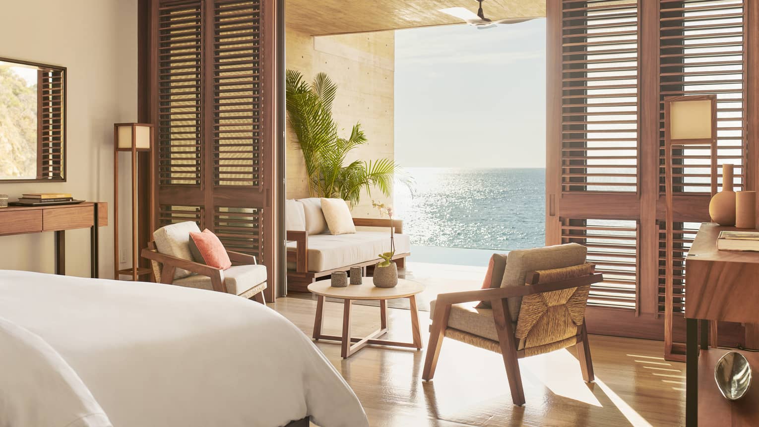 A serene bedroom with wooden accents, featuring a seating area that opens to a terrace overlooking the ocean. Soft natural light fills the room, highlighting the warm, earthy decor.
