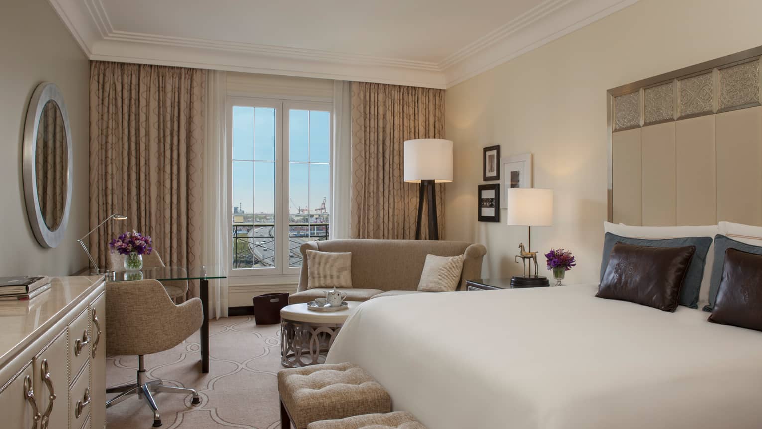 City-View Room with clean white decor, bed with tall padded headboard with silver details, brown pillows, plush benches, chairs