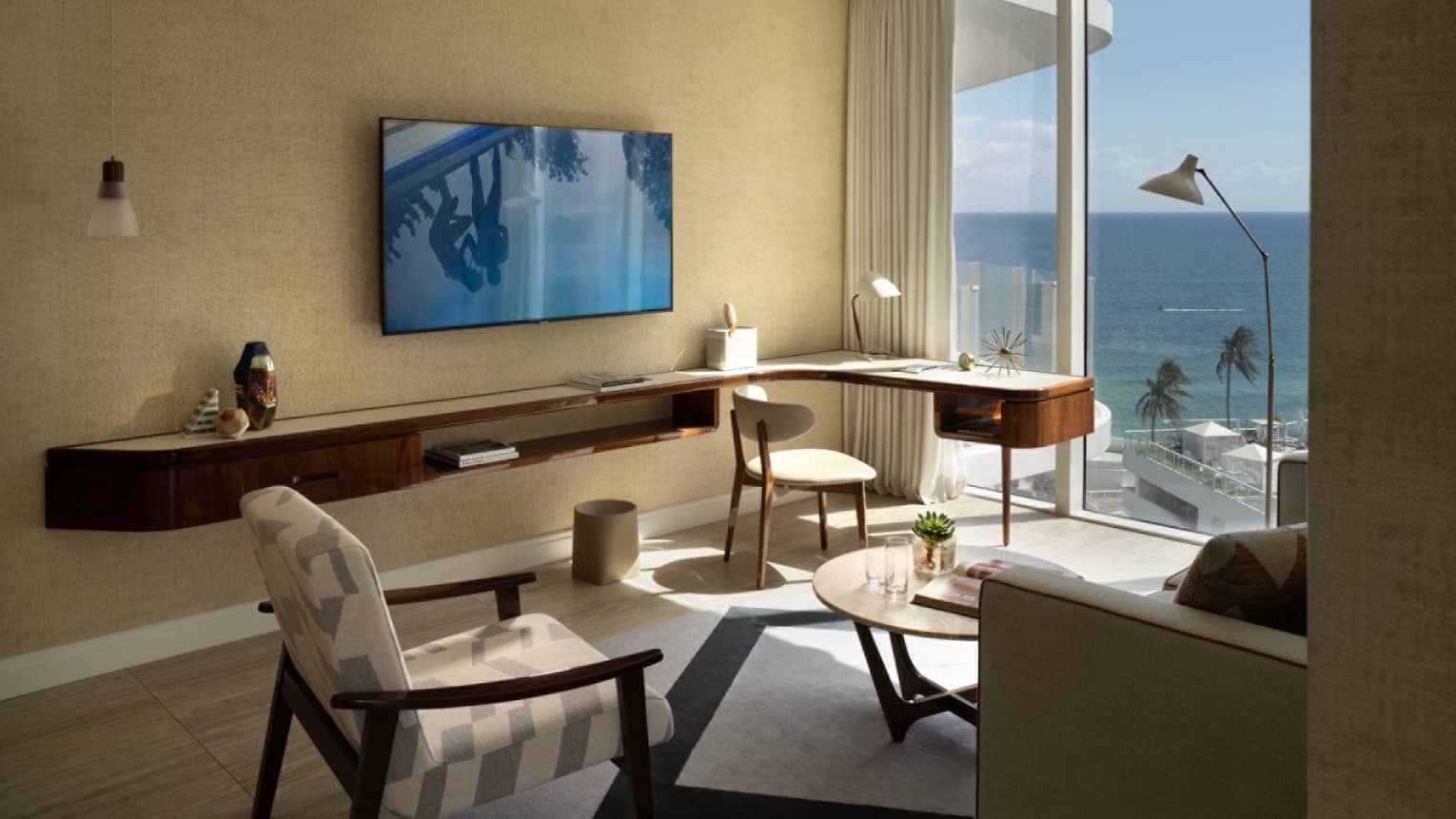 A hotel room featuring a large built-in wall shelf and desk, a sitting area and a wall of windows overlooking ocean