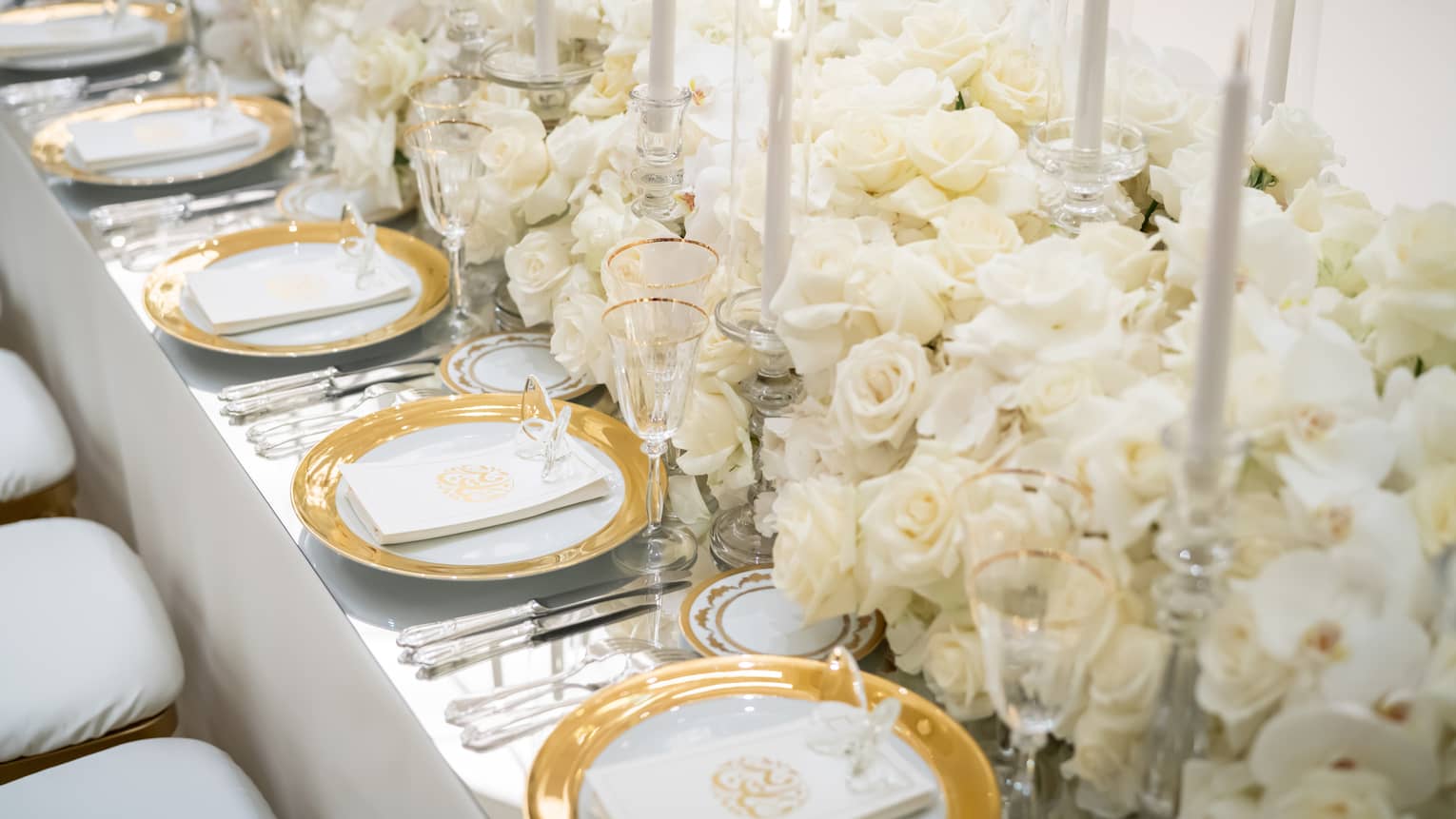 Table filled with white flowers and candles in the centre with a row of white, gold-rimmed plates on either side