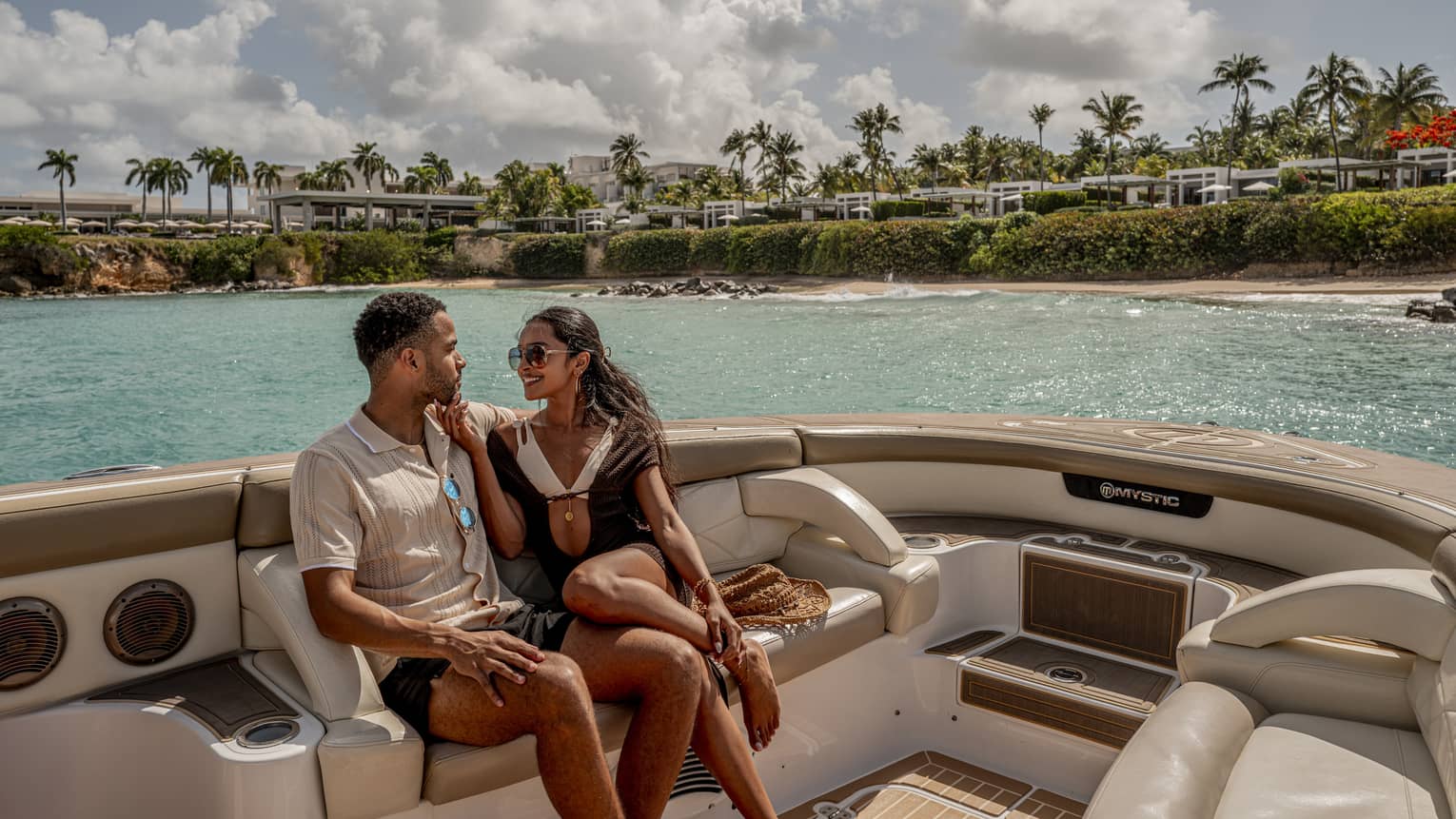 Two people sit on luxury yacht at sea, with palm trees and resort in the background